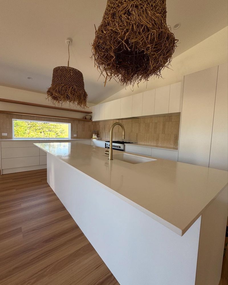 Open-plan kitchen with a white island bench and woven pendant lights in a new home at Korora